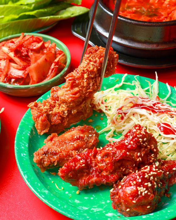 คีบไก่ทอดกรอบขึ้นจากจาน ข้างไก่ซอสเผ็ดโรยงา กะหล่ำปลีราดซอส และกิมจิ