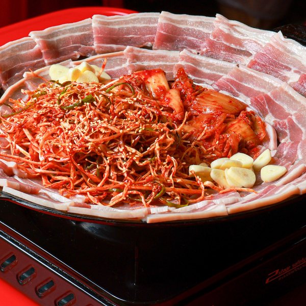 หมูสามชั้นเรียงรอบกระทะ กิมจิกับผักคลุกซอสแดงโรยงาและกระเทียมตรงกลาง วางบนเตาแก๊สพร้อมย่างที่โต๊ะ