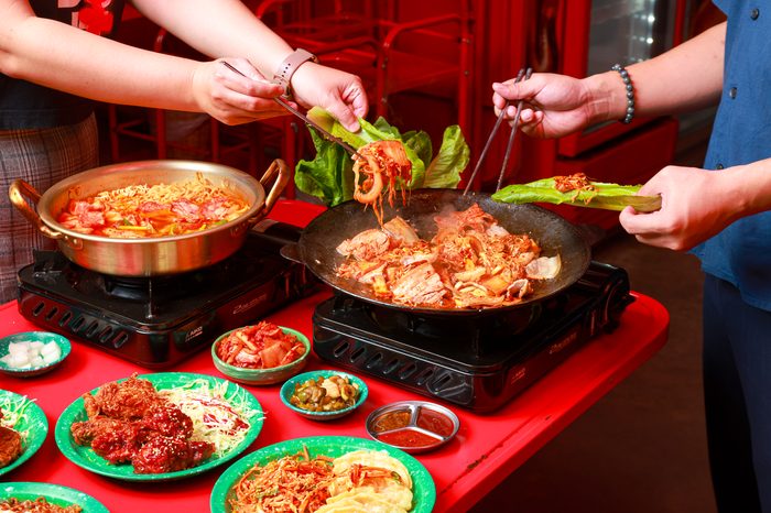 หลายมือช่วยกันคีบหมูย่างคู่กิมจิจากกระทะมาห่อผักกาด ข้างหม้อบูเดจีเก ไก่ทอด และเครื่องเคียงบนโต๊ะแดง