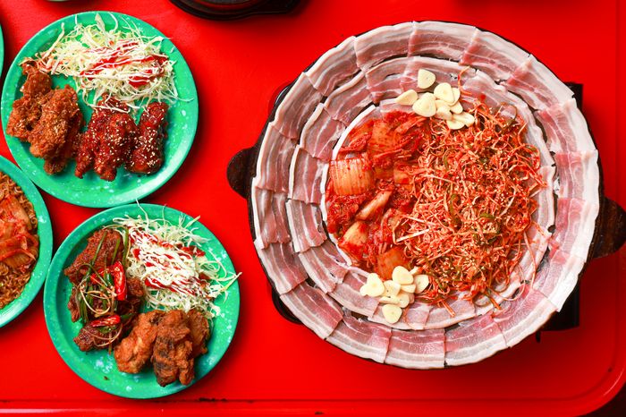 หมูย่างคู่กิมจิบนกระทะร้อน หมูสามชั้นวางรอบกระทะ กิมจิตรงกลาง คู่กับไก่ทอดเกาหลี 2 แบบ บนโต๊ะแดง